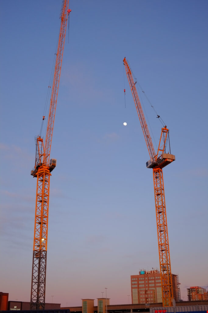 Two cranes in front of the moon appearing in the still blue sky. The crane on the right is positioned so that it tries to grab the moon.