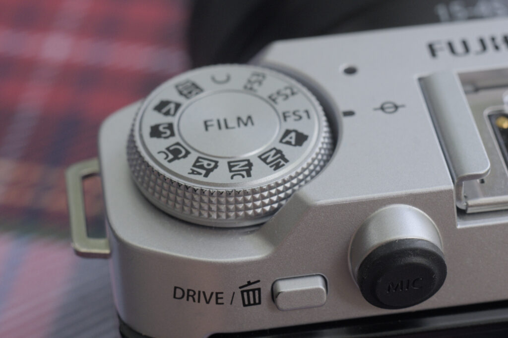 The film simulation dial of the X-M5. It has various symbols with letters denoting specific film simulations CC for Classic Chrome or V for Velvia. Also in some positions C, FS1, FS2 and FS3. The center of the dial is labelled FILM. Just below is a button labelled DRIVE with a trashcan icon, and there is the covered mic input.