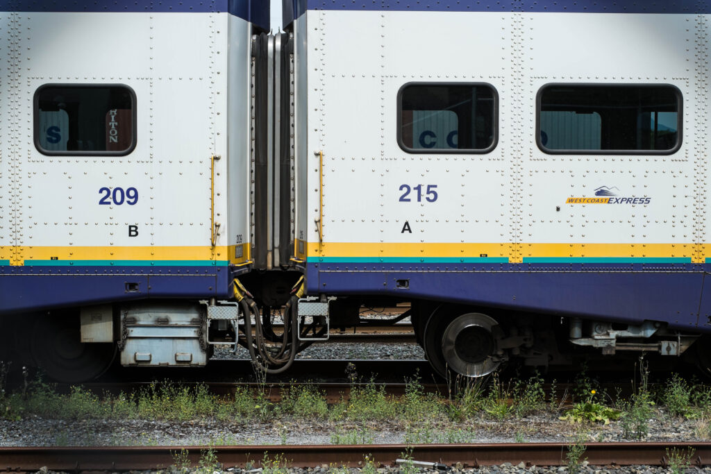 The end of two white suburban train cars connected. With blue stripe at the top and bottom, and the Westcoast Express logo. The train wheels are clearly visible as well as weed growing around the track and in the foreground part of the other empty track.