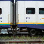 The end of two white suburban train cars connected. With blue stripe at the top and bottom, and the Westcoast Express logo. The train wheels are clearly visible as well as weed growing around the track and in the foreground part of the other empty track.