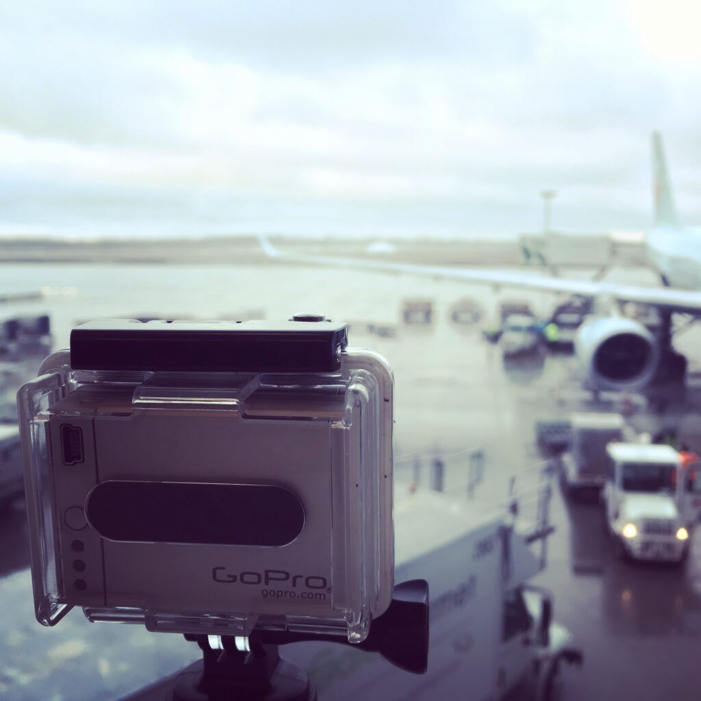 I GoPro camera seens from the back, facing the outside of an airport terminal. Visible in the background, out of focus, is the wing of a jetliner and various service vehicles.